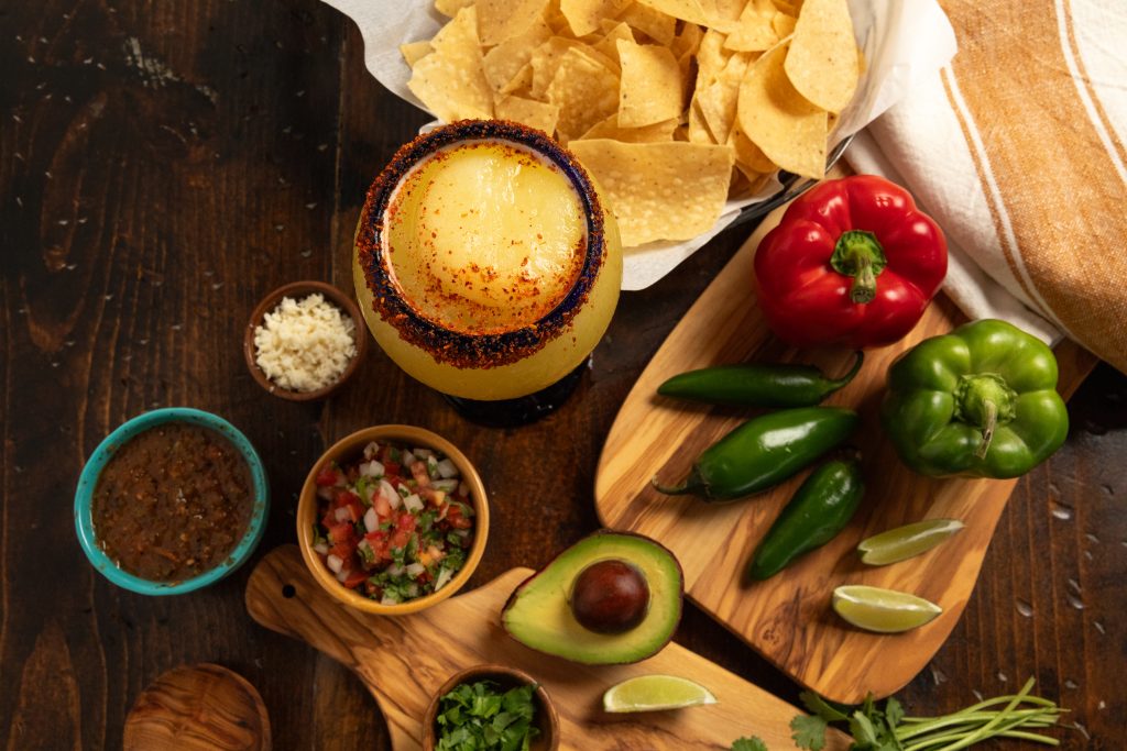 Wooden table holding spices, bell pepper, jalapeños, avocado, chips and margarita.