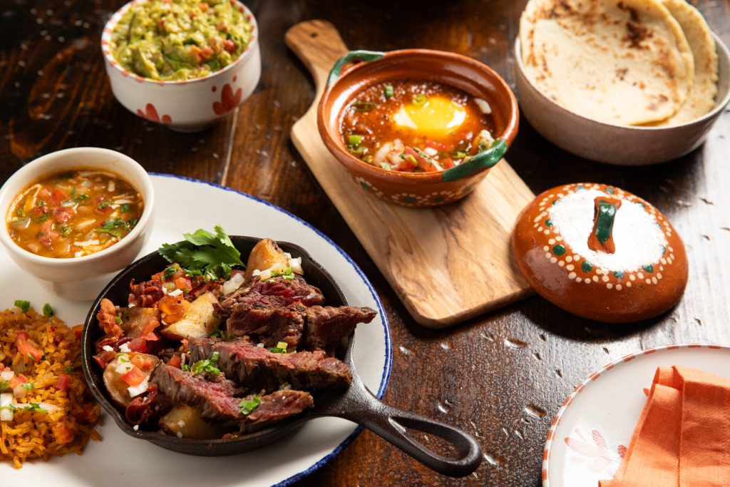 Wooden table with ceramic bowls with guacamole, fajitas, potatoes, rice and beans. Separate bowl with egg and red sauce and tortillas.