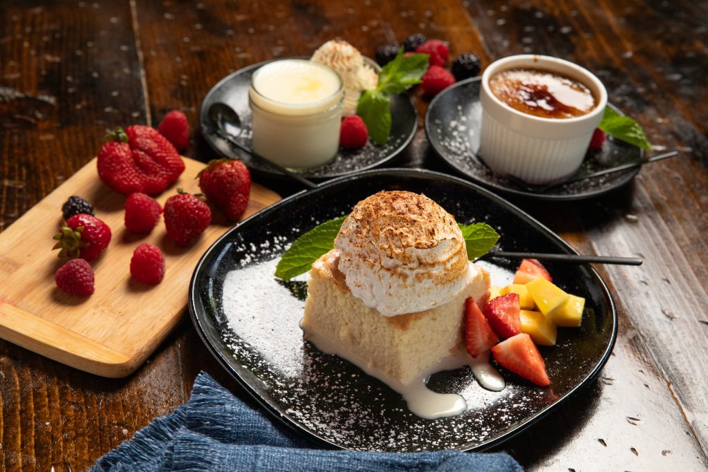 Table set with black plate holding tres leches, flan and pina colada desserts. Garnished with raspberries.