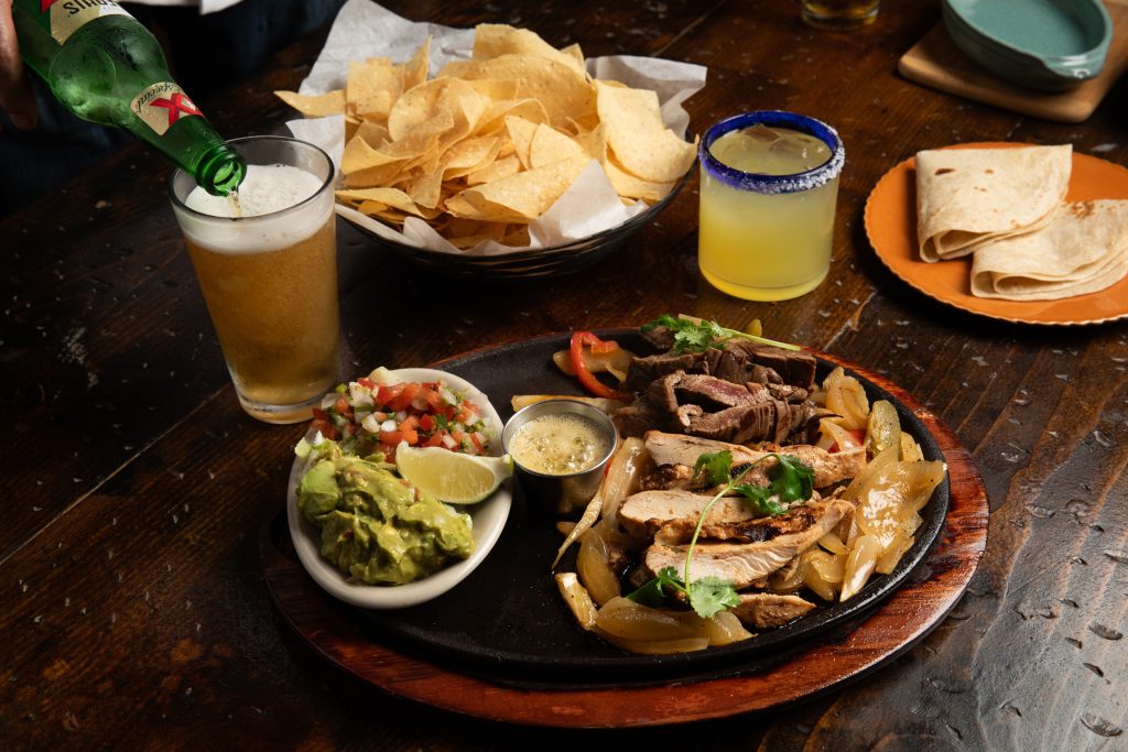 Wooden table with margarita, chips, flour tortillas and fajita with guacamole. Beer is being poured.