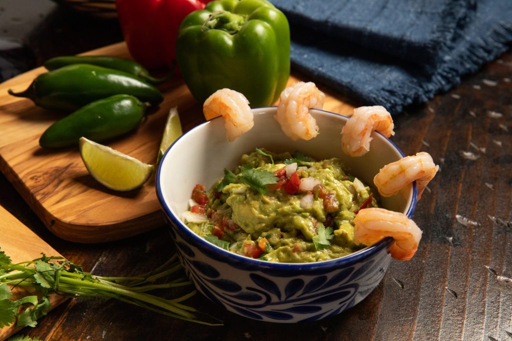 Bowl of guacamole with shrimp on wooden table with blue napkin. Also on table is jalapeno, lime slice and bell pepper