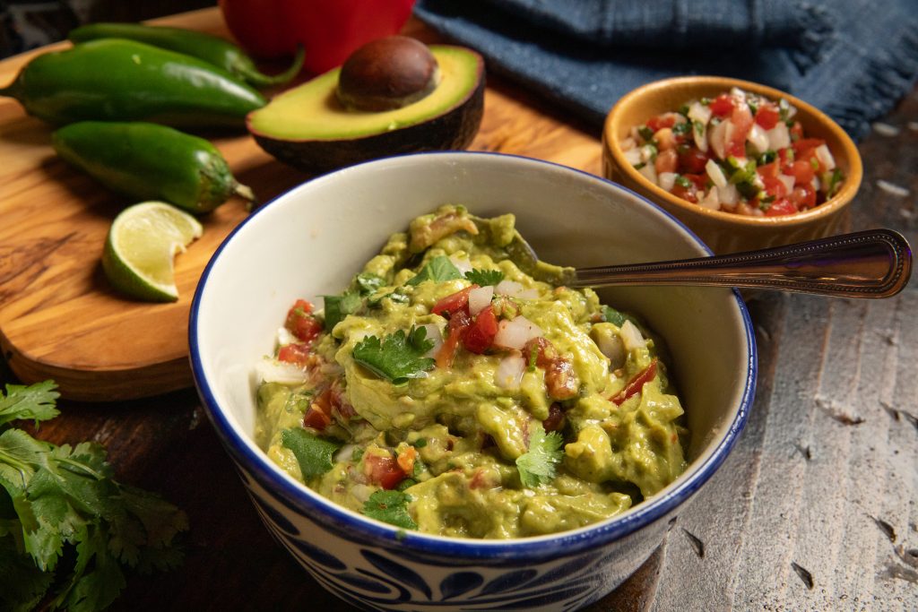 Wooden table with guacamole, chips and salsa along with peppers, onions, limes and jalapeños.