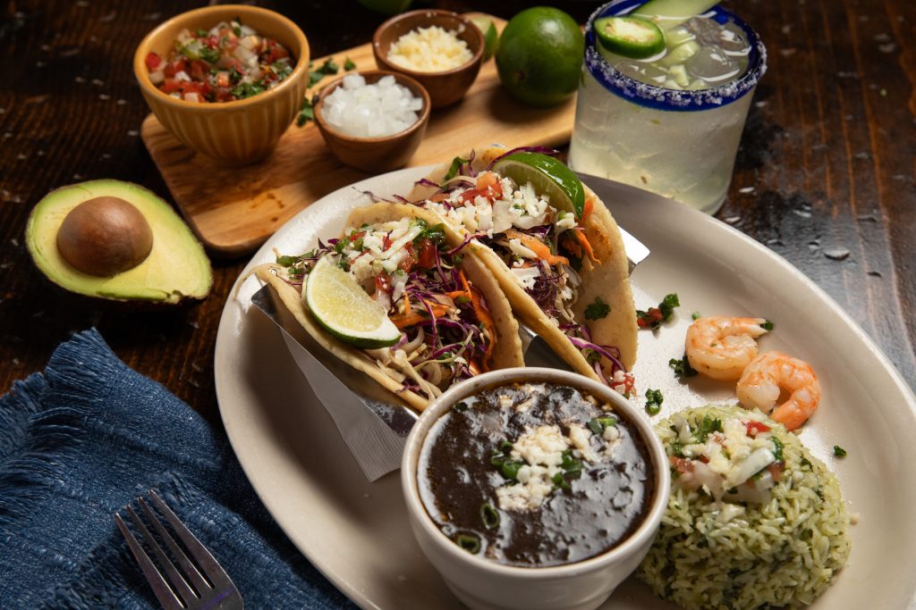 Plate of shrimp tacos with black beans and rice together with jalapeno margarita. On the side are spices and avocado.