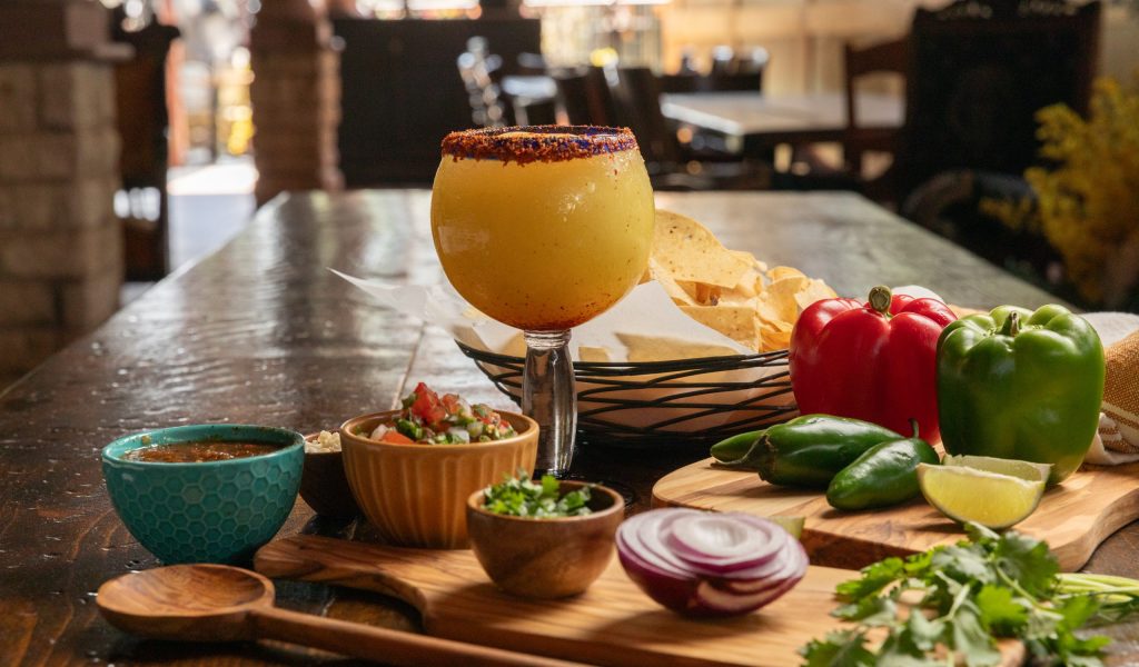 Table set with margarita chips together with onion, peppers, jalapeños on wooden tray with wooden spoon.