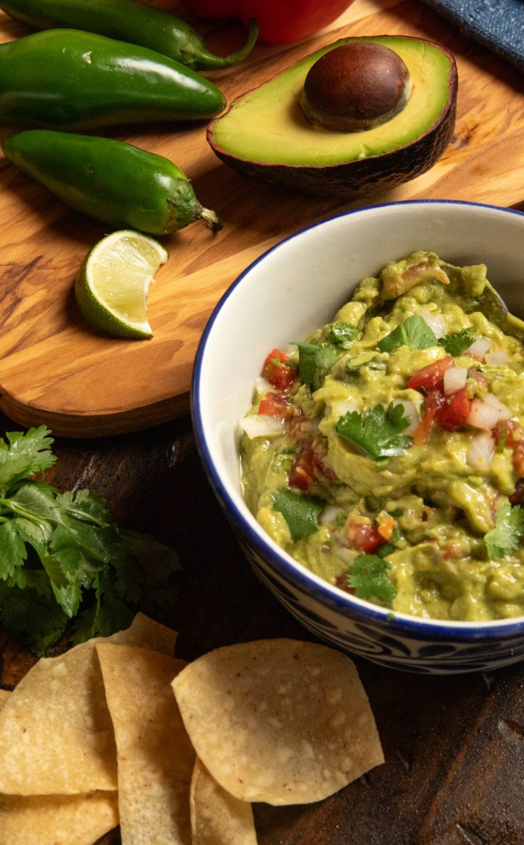 Wooden table with guacamole, chips and salsa along with peppers, onions, limes and jalapeños.