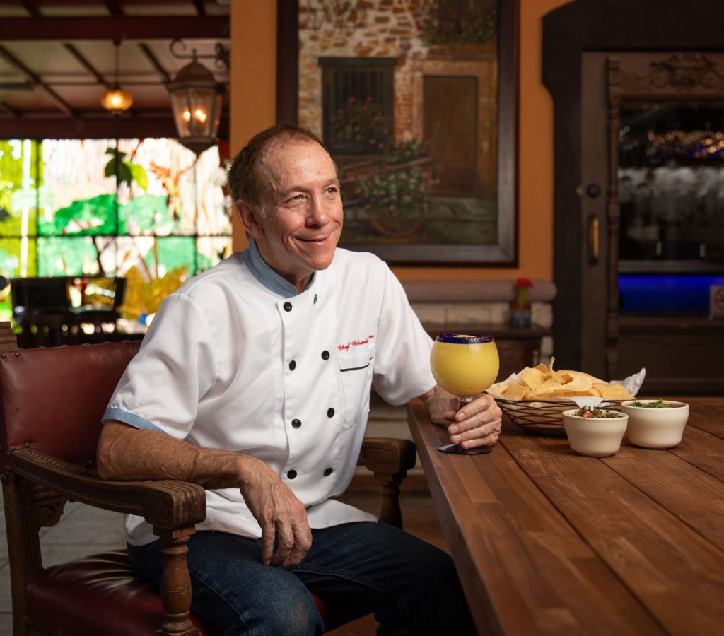 Chef Charlie sitting at table with a margarita in hand. Colored glass window behind chef.