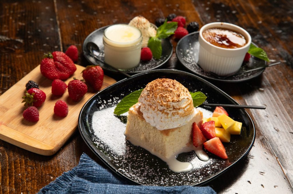 Table set with black plate holding tres leches, flan and pina colada desserts. Garnished with raspberries.