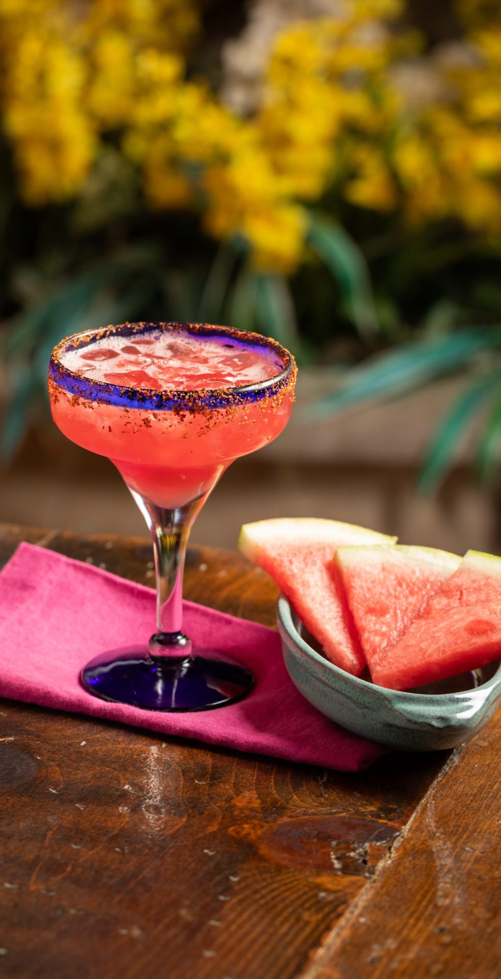 Watermelon Margarita place on table with pink napkin, watermelon slices with yellow flowers in background