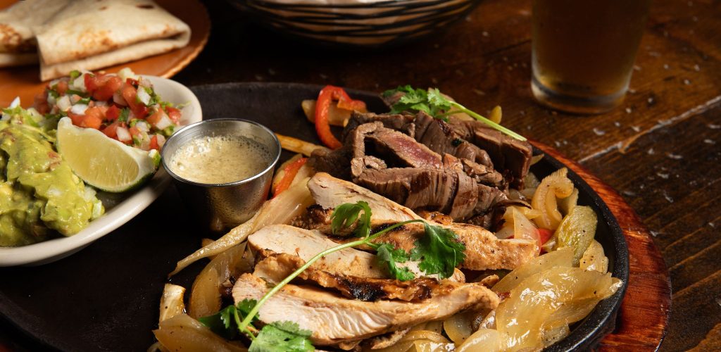 Chicken and beef fajitas platter on wooden table with tortillas and guacamole