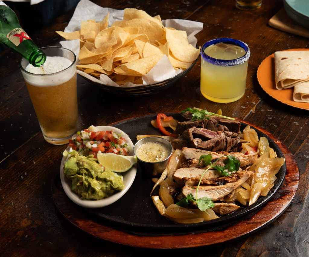 Wooden table with margarita, chips, flour tortillas and fajita with guacamole. Beer is being poured.